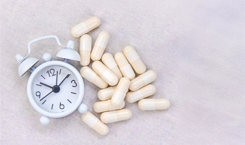 Natural melatonin sleep supplement capsules arranged next to white alarm clock on beige fabric background