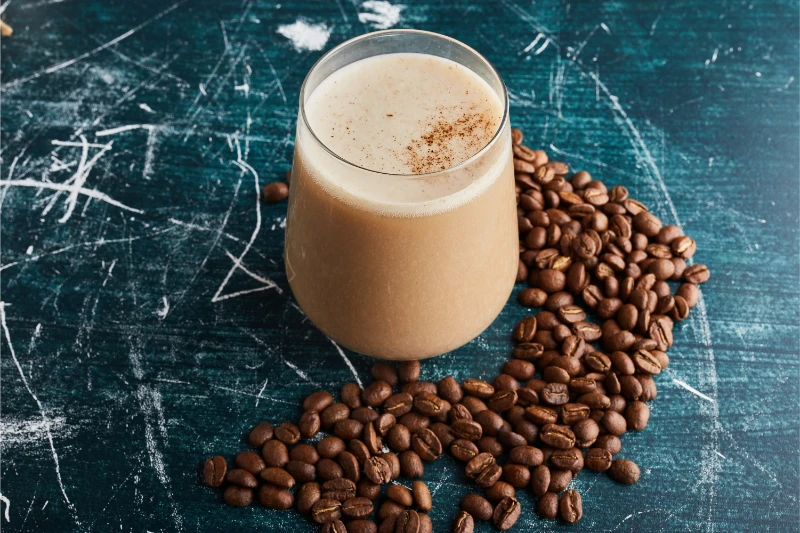 Creamy iced coffee protein shake in a glass surrounded by coffee beans on a teal wooden table