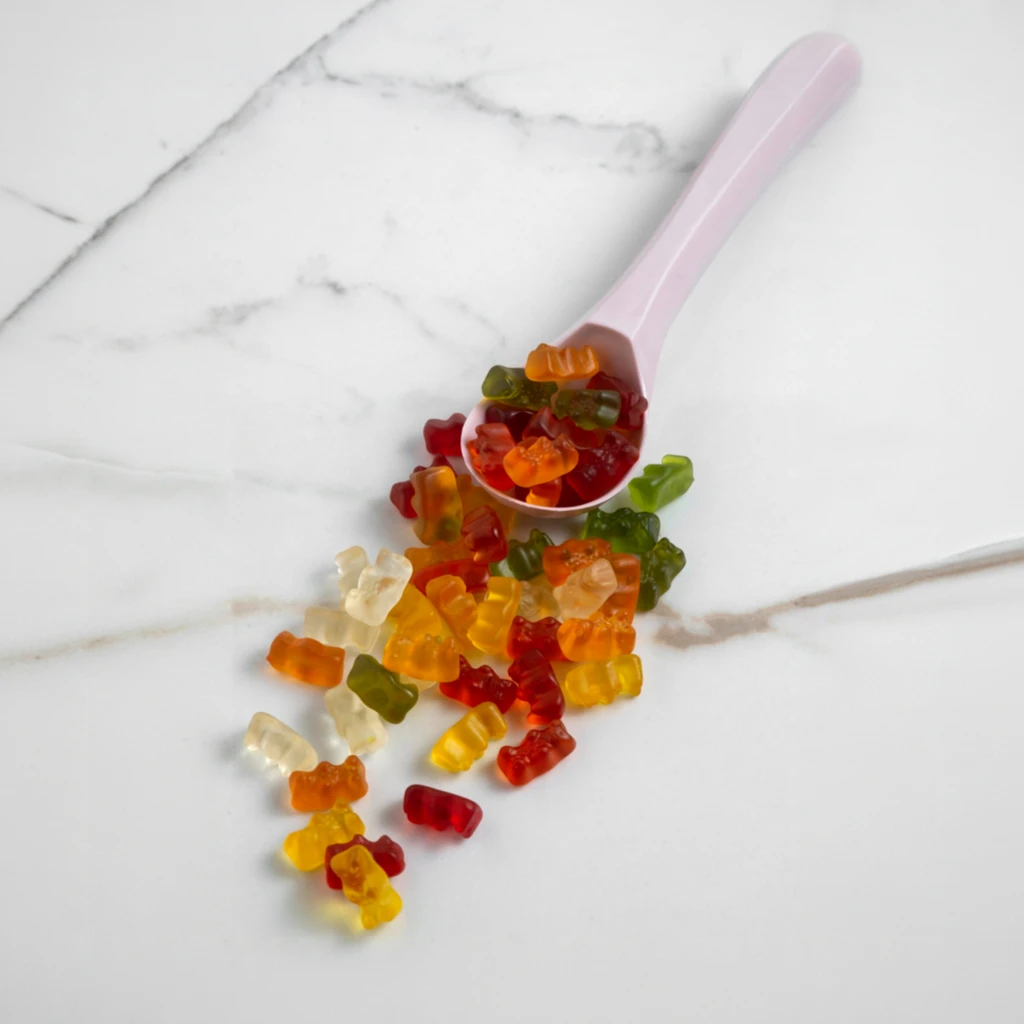 Colourful creatine gummies in red, orange, yellow and green spilling from a pink measuring spoon onto a white marble surface